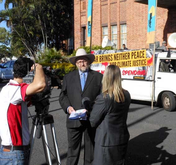 Bryan Law wearing a white hat and suit in his theatre costume of 'peace preacher' being interviewed by a news reporter. A cameraman is filming him. There is a van in the background with anti-war slogan.