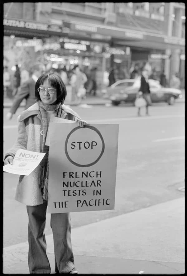 Woman holding sign and handing out leaflets on the street. The sign says Stop French Nuclear Tests in the Pacific.