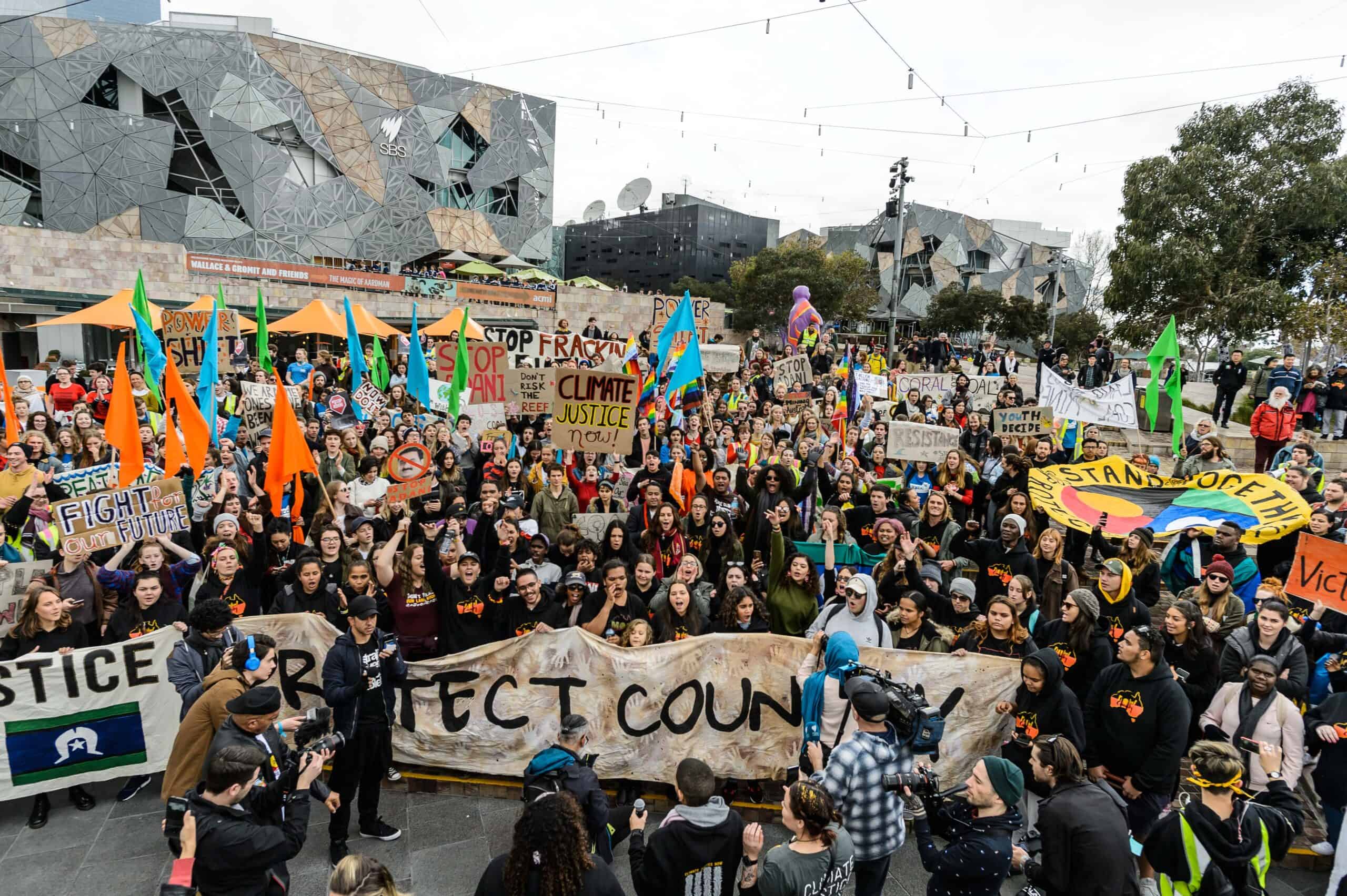 Photo of a climate protest in Melbourne's Federation Square. Protesters hold various placards and banners with climate slogans.