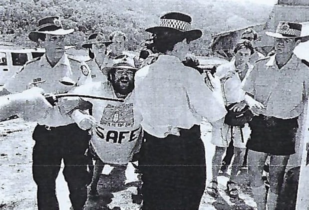 Black and white photo of a man being arrested by three police at a protest. The man being arrested is wearing a t-shirt that says Safe.