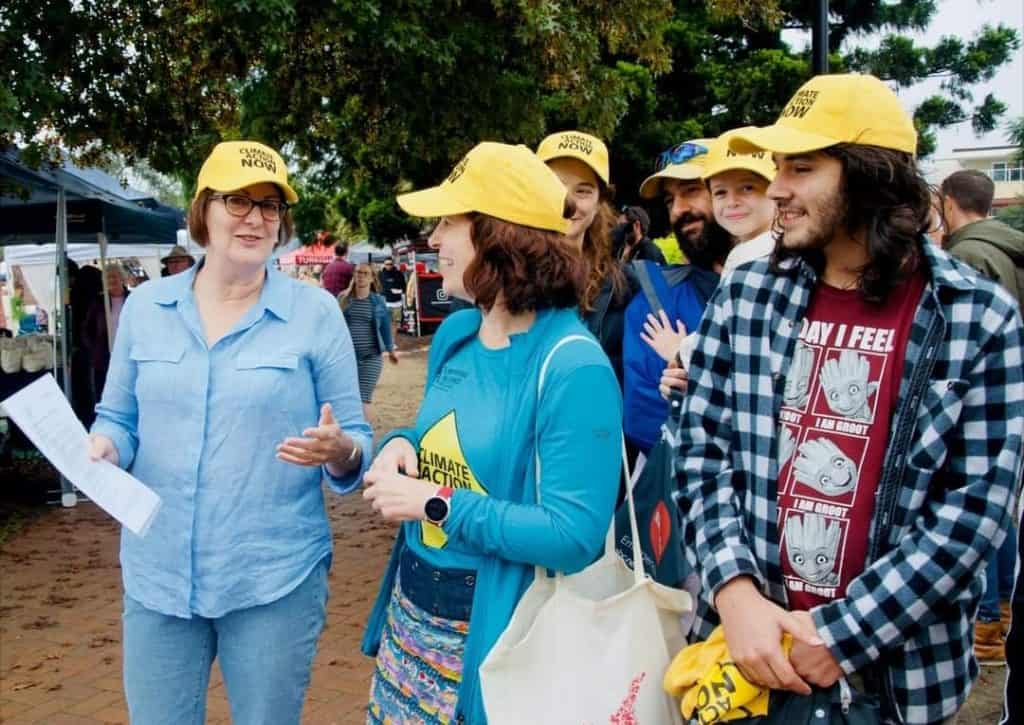 a group of people outside wearing yellow caps talking together at a market. One woman's tshirt reads Climate Action Now.