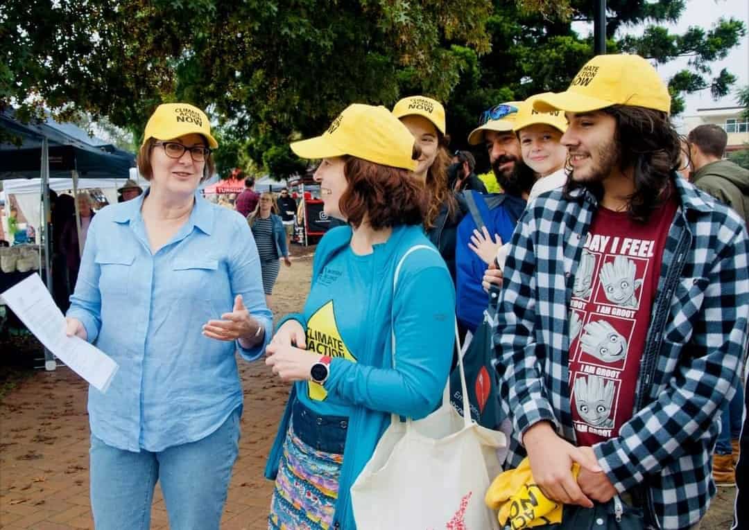 a group of people outside wearing yellow caps talking together at a market. One woman's tshirt reads Climate Action Now.|man knocking on door knocker of a black door. He is wearing a tshirt that reads Stop Coal - Stop Adani Take Action