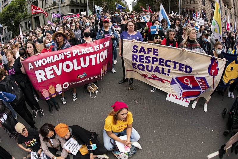 Photograph of a rally showing several women holding banners including: 'A Women's Place is in the Revolution' and 'Safe