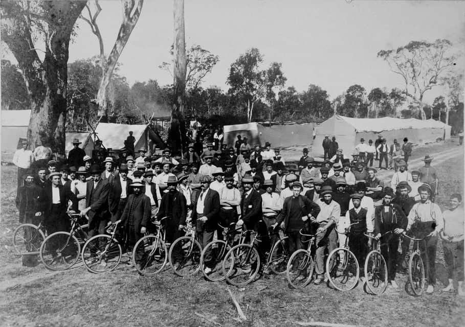 A large group of men gathered outside amongst gum trees and strike camp with bikes.