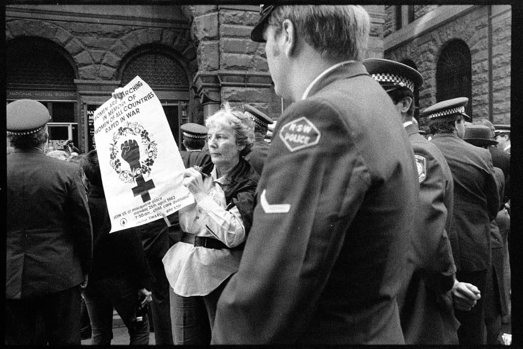 Woman holding sign about anti-conscription with policeman standing in front of her.