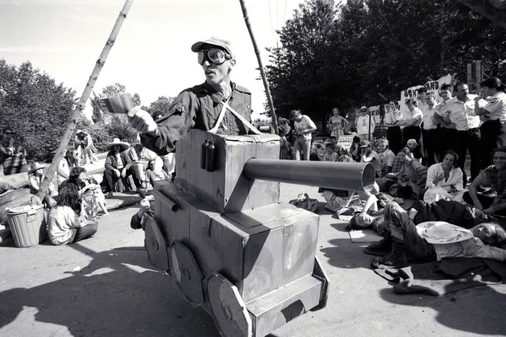 Man dressed up in army gear in a cardboard box army tank in front of a group of protestors. He is part of the Members of the Sydney Peace Squadron performed street theatre throughout the AIDEX blockade in Canberra, Australia in 1991.
