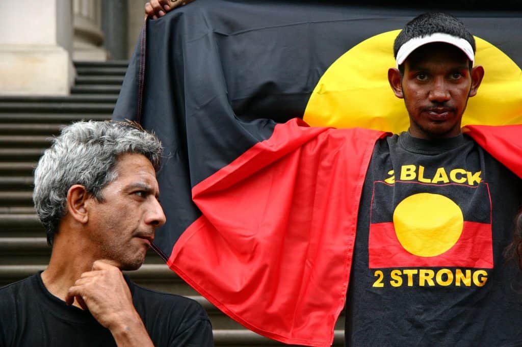 Man with Aboriginal flag draped around his shoulders wearing tshirt with Australian Aboriginal flag that says Black 2 strong