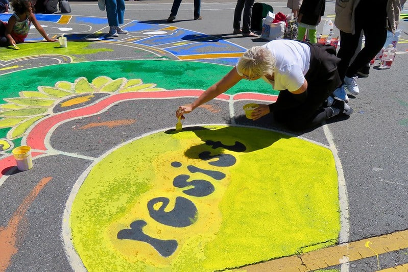 woman kneeling on the ground drawing in chalk the word resist inside yellow circle for a climate action in San Francisco