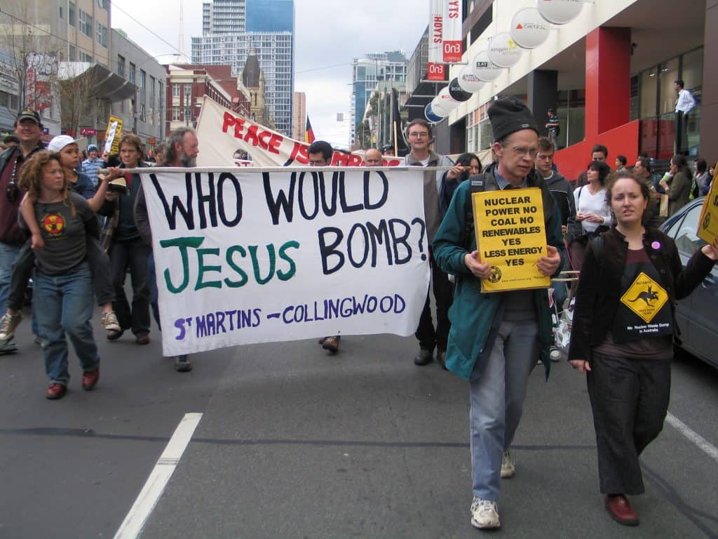 Protestors holding banners walking down street. One banner says Who would Jesus bomb?