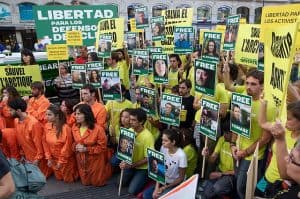 Protestors, some wearing yellow Greenpeace shirts, some in prison orange, pose for a photo holding signs in Spanish, and photographs of the activists detained by Russia known as the Arctic 30.