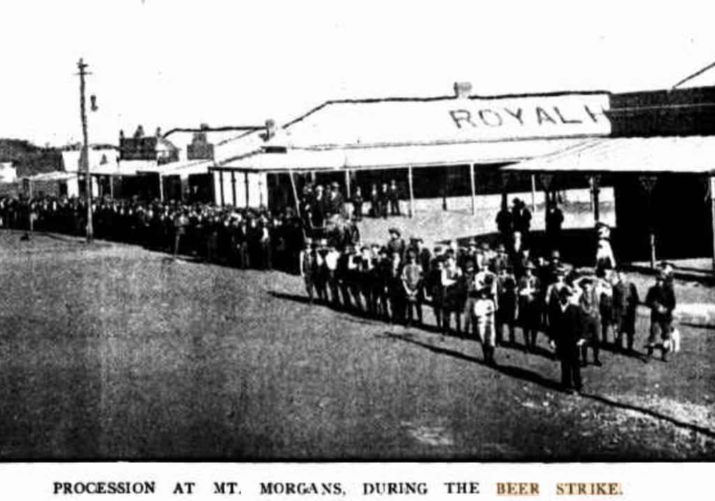 an old black and white photo of a procession of people walking along a street outside a hotel with text on their roof that reads Royal Hotel