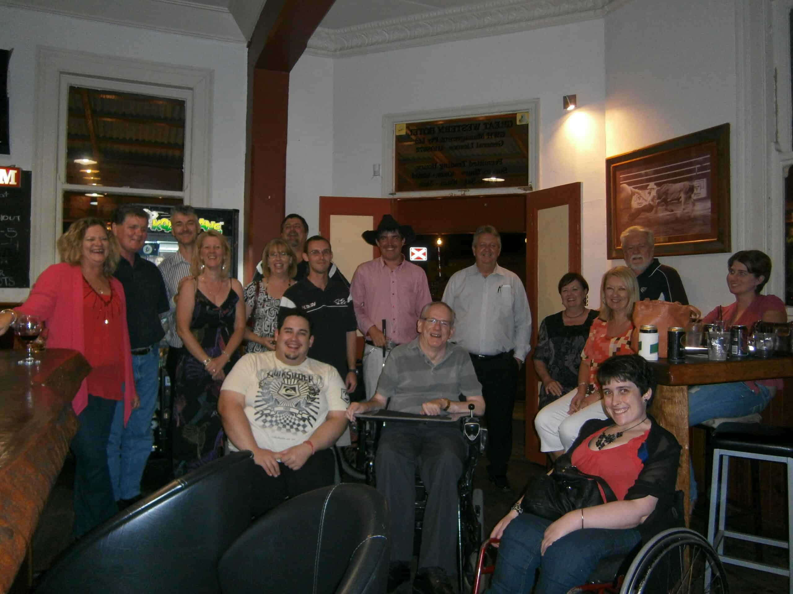 A group of people at the pub smiling for the camera. The people have different disabilities including wheelchair users and blind people.