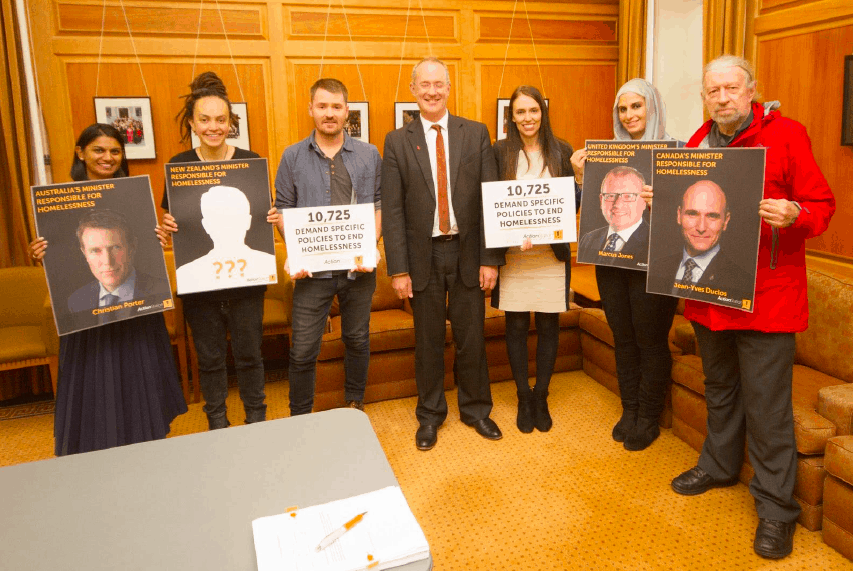 A group of community members and politicians hold placards including '10,725 demand specific policies to address homelessness'.