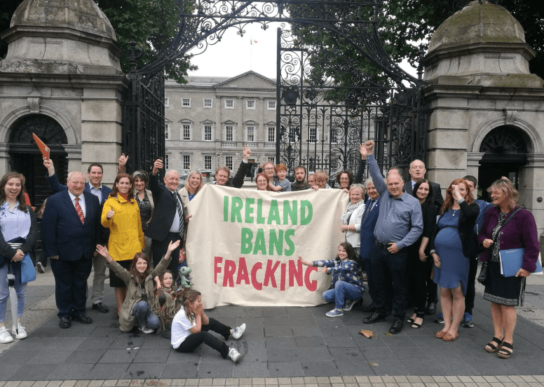 a group of people standing outside a large building with a metal gate holding a banner that reads Ireland bans fracking.