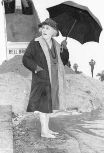 A 89 year old woman wearing a hat, knee length coat and dress stands in bare feet in the water of a bay holding an umbrella standing in front of a pile of dumped sand being tipped out of a truck