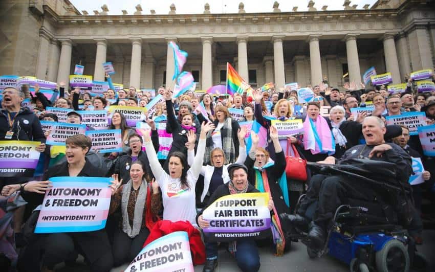 A crowd stands in front of the Victorian State Parliament. They are holding placards in the colours of the trans flag (pink, blue and white).