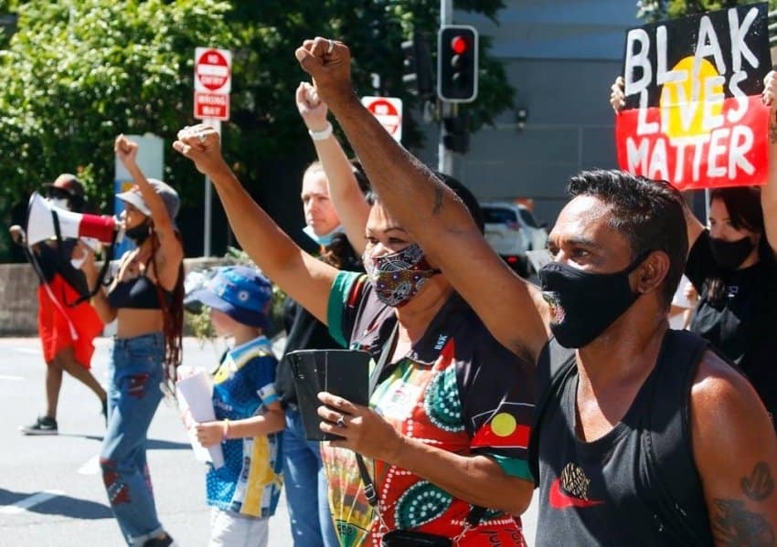 a group of people protesting on the street wearing face masks and holding their fists in the air. There is a banner in the back reading Black Lives Matter. The person at the front wearing an Australian Aboriginal flag is holding their phone.