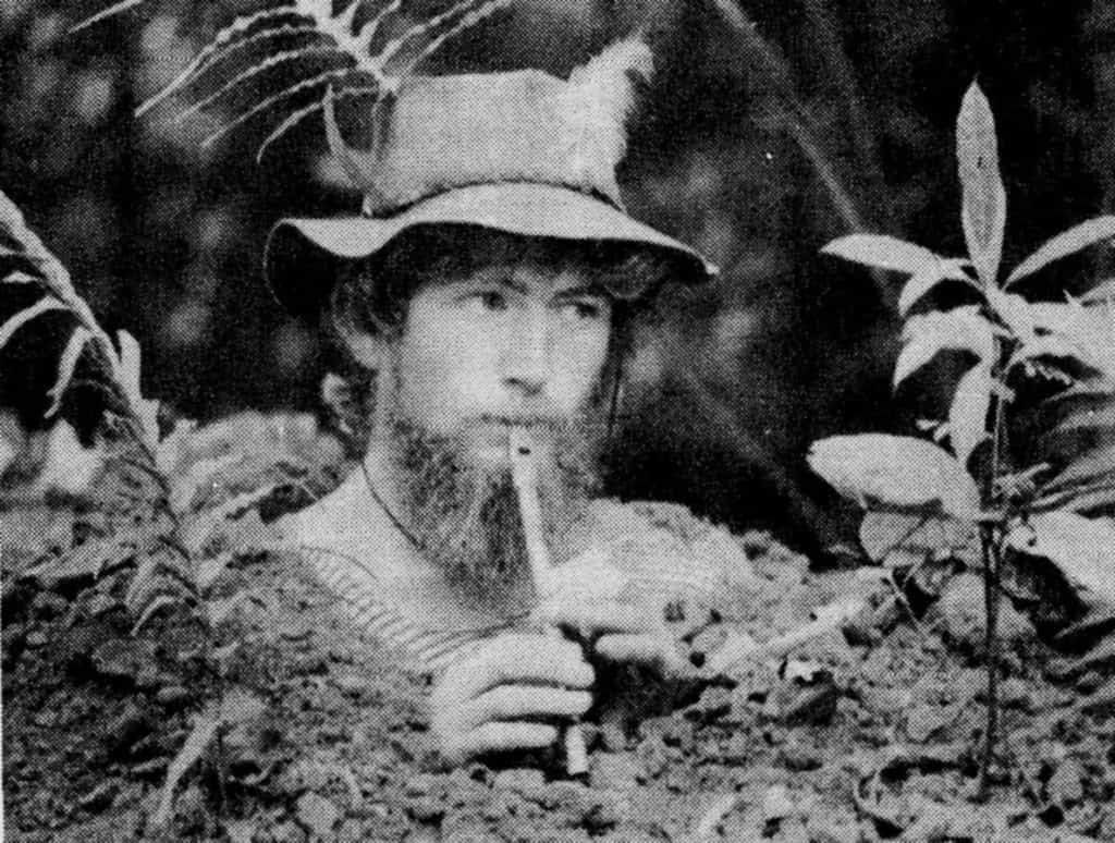 man buried in the earth in soil with his head poking out. He is wearing a hat and playing a recorder.