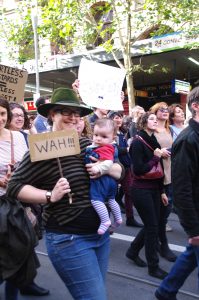 Protesters march down the street. A smiling woman holds a baby with one arm, in the other hand she holds a small placard that reads 'WAH!!!'