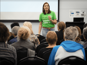 A young woman stands in front of a seated group of people. She is wearing a green tshirt that reads 'We are a powerful force for nature'
