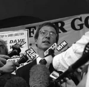 A man wearing glasses, Dave Burgess, speaking to the media. Hands with microphones are held in front of him. There is a banner in the background.