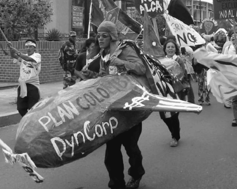 A person walking down in the street at a protest in Quito in Ecuador. The protestor is dressed up as a pilot with a cap and goggles and is wearing a large cardboard plane around his body. On the plane it reads 'Plan Colombi DynCorp'. There are other protestors behind them holding flags and signs.
