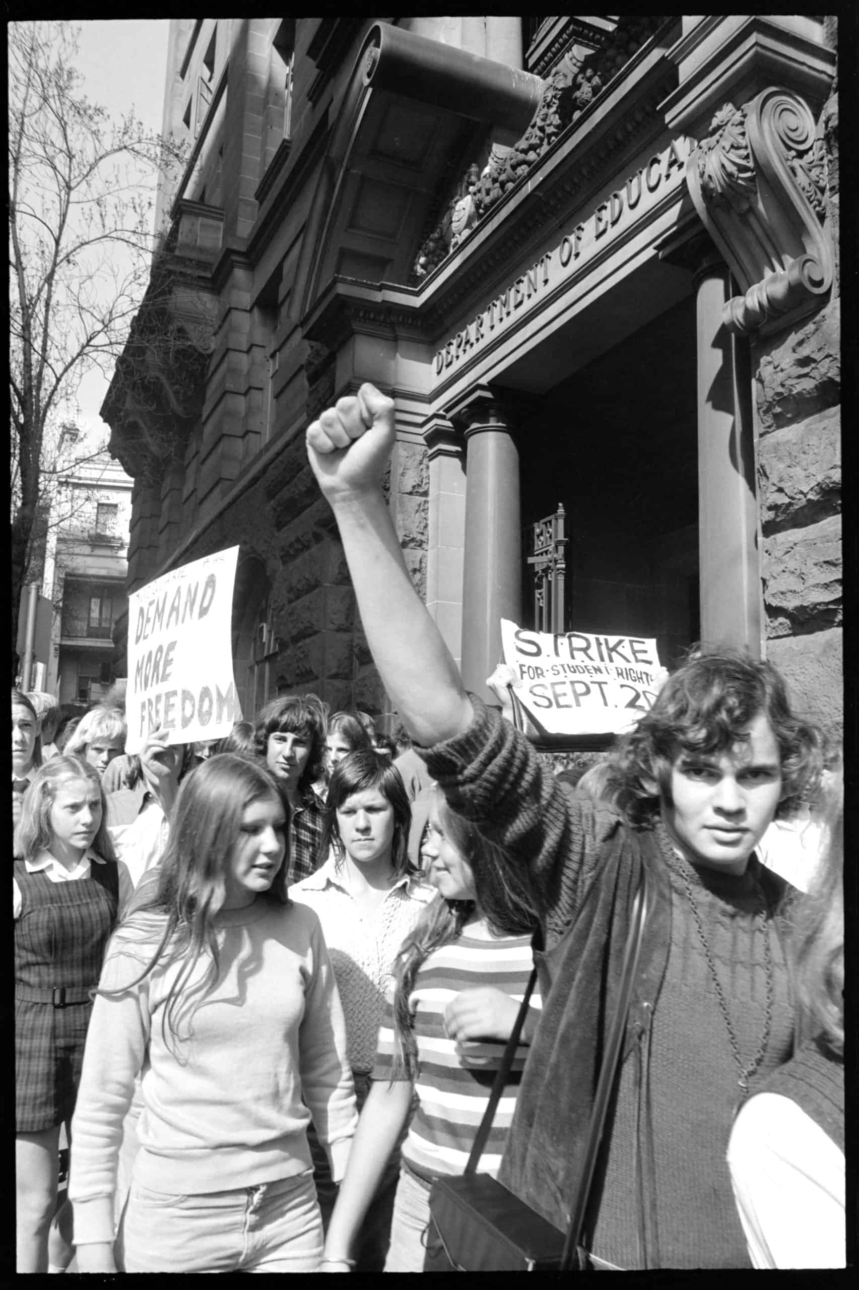 Students walking along street outside an ornate building that reads Department of Education over the doorway. Some students are holding placards. The student at the front has their fist in the air.