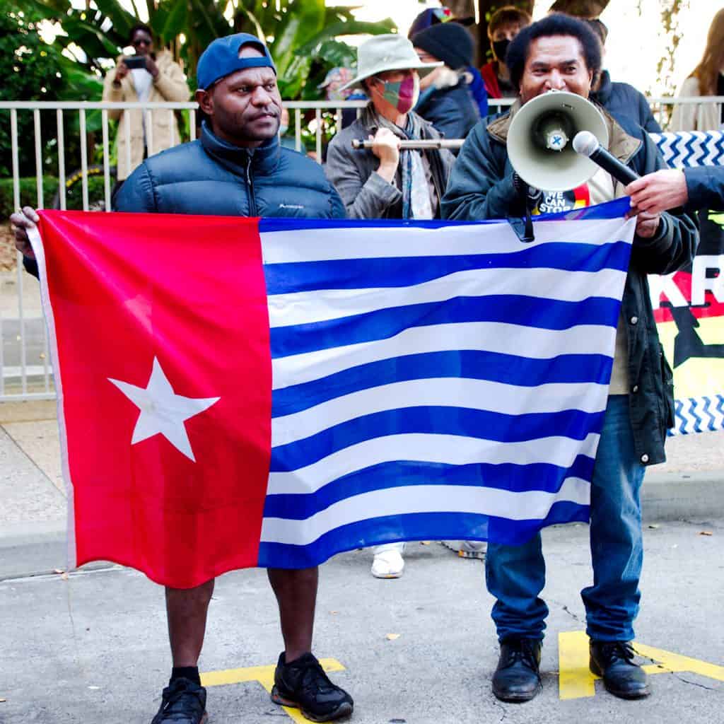 3 protestors holding up the West Papua flag. One protestor is holding a megaphone