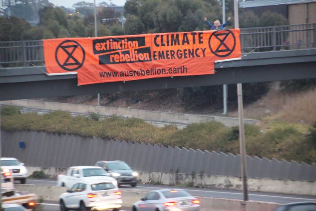 Orange banner draped over pedestrian bridge over freeway full of cars. The banner says Extinction Rebellion Climate Emergency.