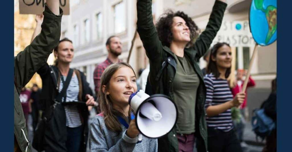 people walking down the street protesting. At the front of the group is a young girl holding a loudspeaker. Next to her is a woman with both hands raised.
