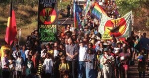 Photograph of protestors at the Jabiluka blockade in 1998. Features banners with 'Uranium, Leave it in the Ground'. The march is led by Mirarr elders and Peter Garrett.