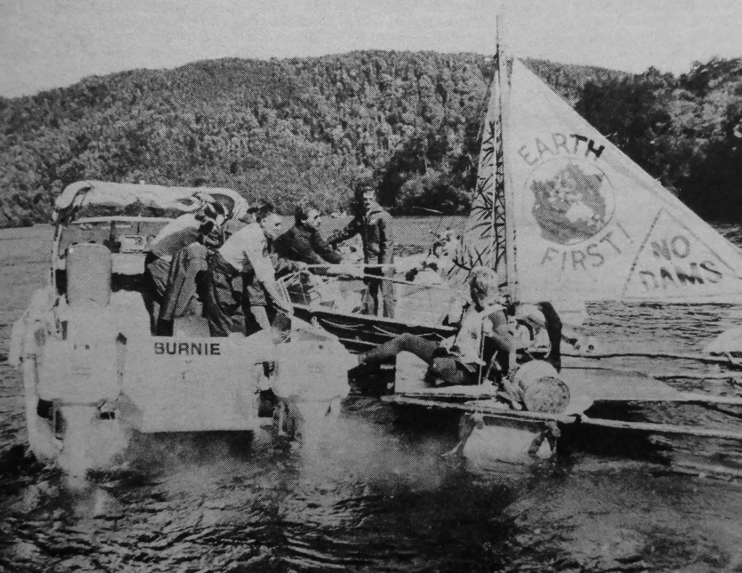 Three boats on a river. Two speedboats with police trying to stop the third sailing boat with protestor. The sail says Earth First and No Dams.