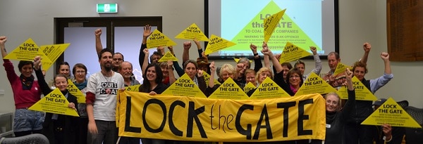 A group of people stand behind a Lock the Gate banner and hold yellow triangles in the air.