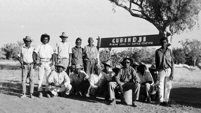 Group of men stand and sit in front of a sign reading 'Gurindji Mining Lease and Cattle Station'