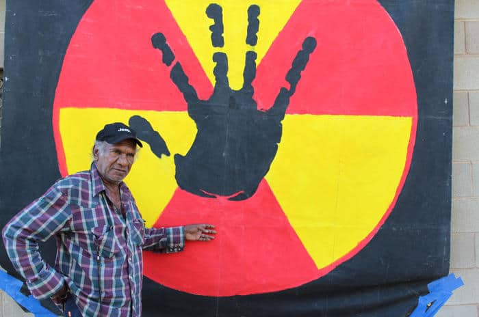 Uncle Kevin Buzzacott stands in front of a large banner depicting a black hand against a red and yellow nuclear symbol.
