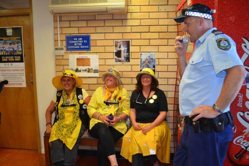 Three women sitting on bench at police station with police man standing on the left. The women are all smiling and wearing yellow hats and black and yellow clothing.