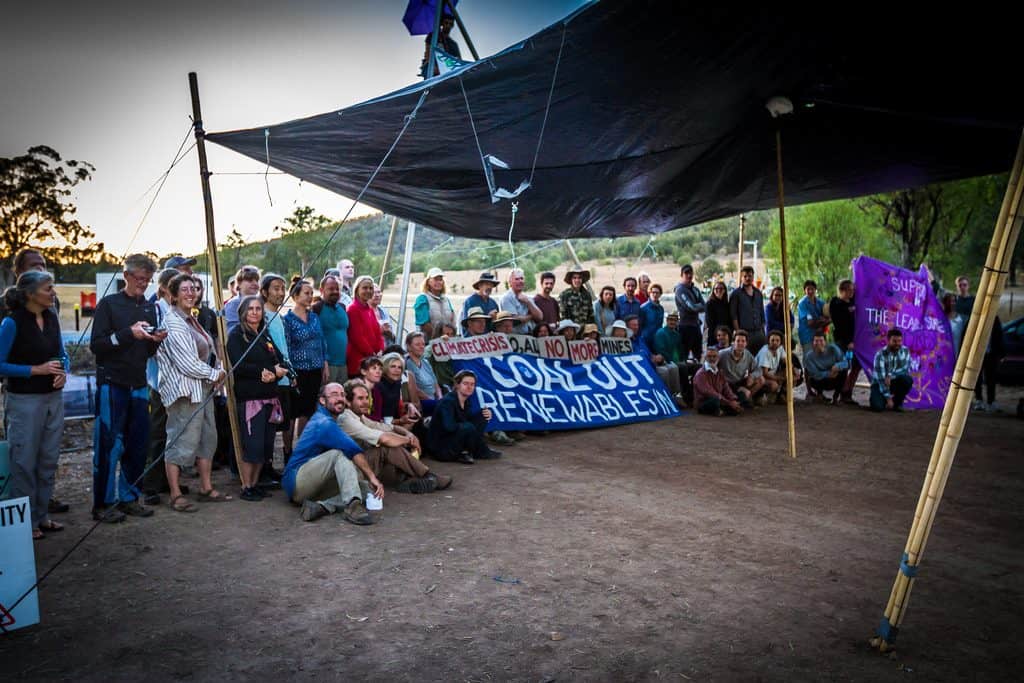 A group of smiling protestors under a tarp shelter. A banner reads 'Coal Out, Renewables In'.