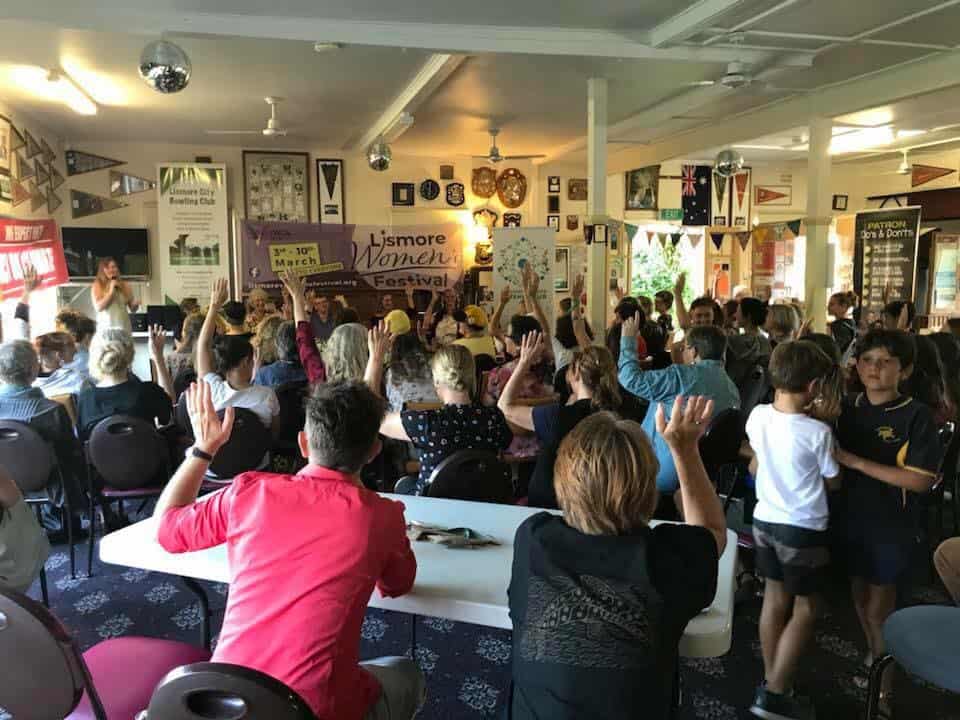 A room full of women at a forum raising their hands, photographed from the back of the room.