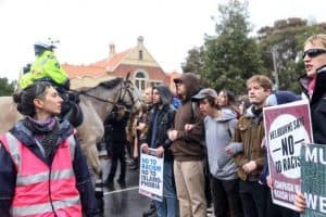 A legal observer in a bright pink vest stands to one side, watching activists holding anti-racist placards. Police on horseback are behind the observer.