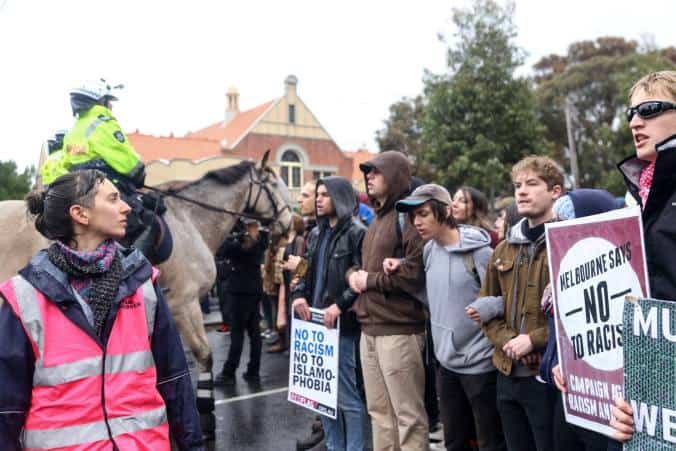 A legal observer in a bright pink vest stands to one side, watching activists holding anti-racist placards. Police on horseback are behind the observer.
