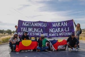 Blockaders sitting on the road with Aboriginal Australian flags and a large banner at the back that reads - Ngiyaningu Maran Yaliwunga Ngara-Li - Our ancestors are always watching.