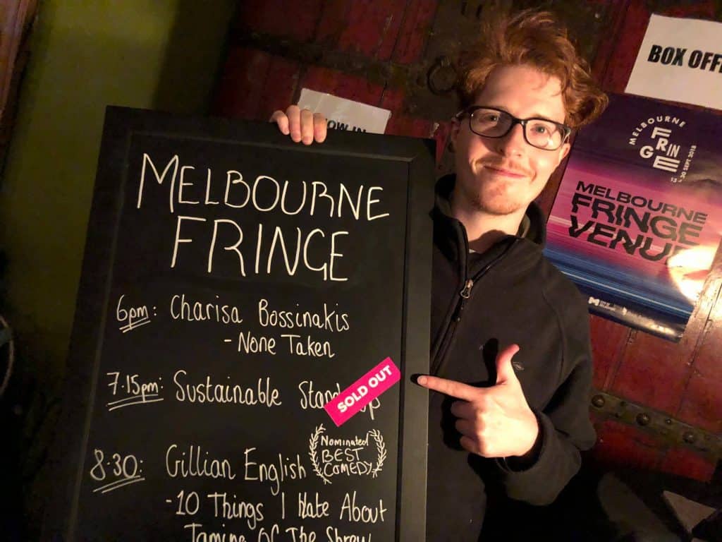 A young man with red hair and wearing glasses points to a blackboard sign titled 'Melbourne Fringe'. His finger points to the 'Sold Out' sticker next to 'Sustainable Standup'.