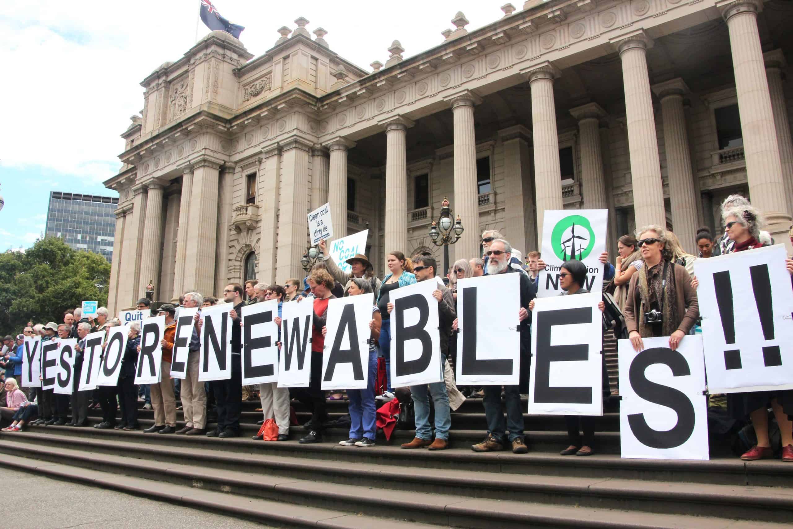 Protestors standing on the steps of the Victorian Parliament building. Each person is holding a poster with a letter that spells out Renewables!!