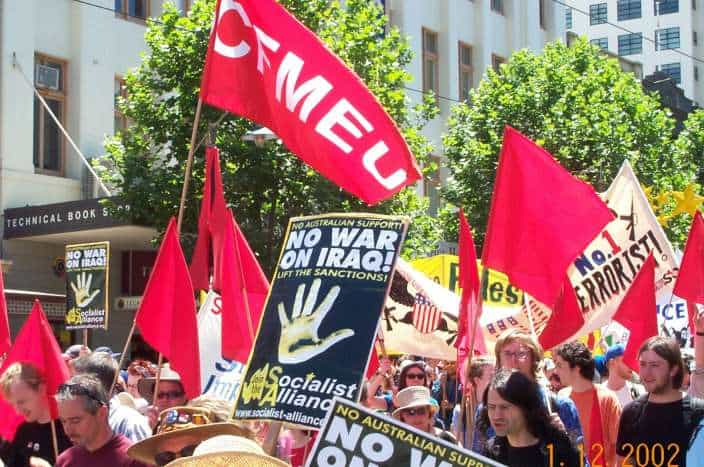 Protestors marching down street. Posters held in the air that say No War on Iraq and a red flag with CFMEU.