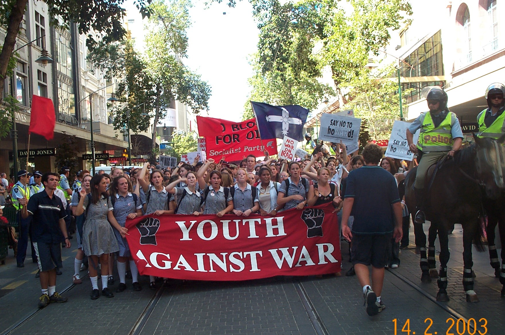 Protestors marching down street holding banners. The banner across the front marchers says Youth Against War