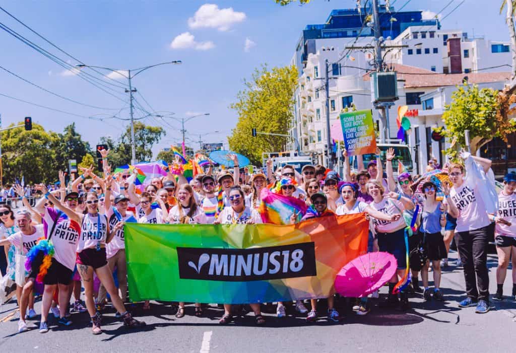 Photograph of a jubilant crowd of young people wearing 'Queer Pride' tshirts, standing behind a rainbow coloured Minus18 banner.