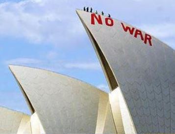 Close up of two Opera House building sails or the triangular parts of the building. On one reads NO WAR in red paint and there are people standing on top of the sail. They are silhouetted against a blue sky with faint white clouds.|A man wearing glasses