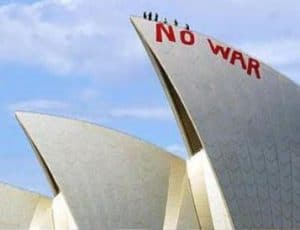 Close up of two Opera House building sails or the triangular parts of the building. On one reads NO WAR in red paint and there are people standing on top of the sail. They are silhouetted against a blue sky with faint white clouds.