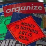 an outdoor post plastered with stickers with the writing organize and money is nothing art is real