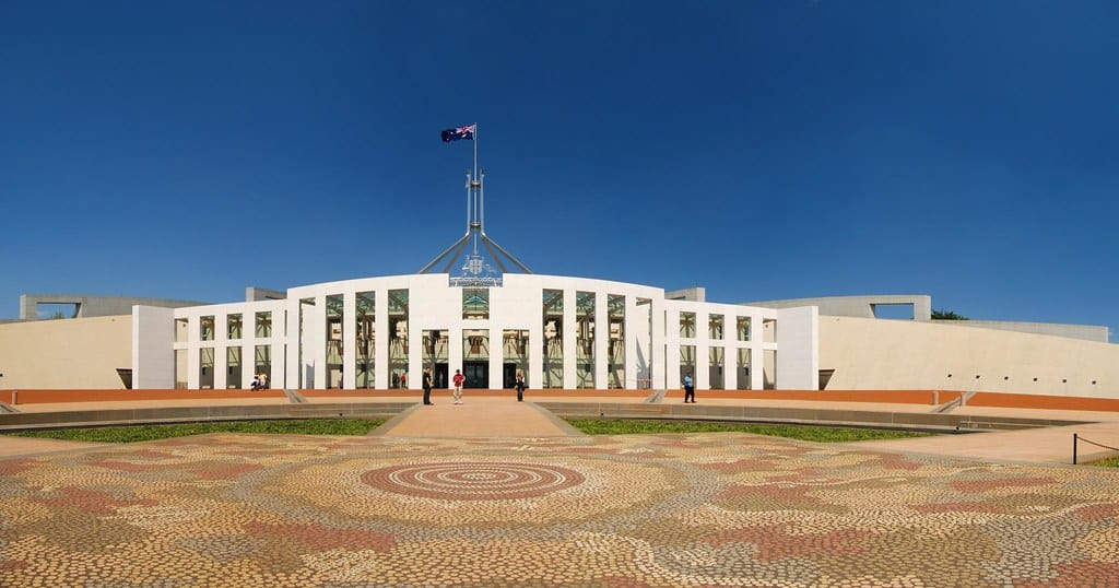 Photograph of the front of Australian Parliament House. The front paving shows a pattern that echoes Aboriginal artwork. The building is white with a flag spier and visible Australian flag blowing in the wind.